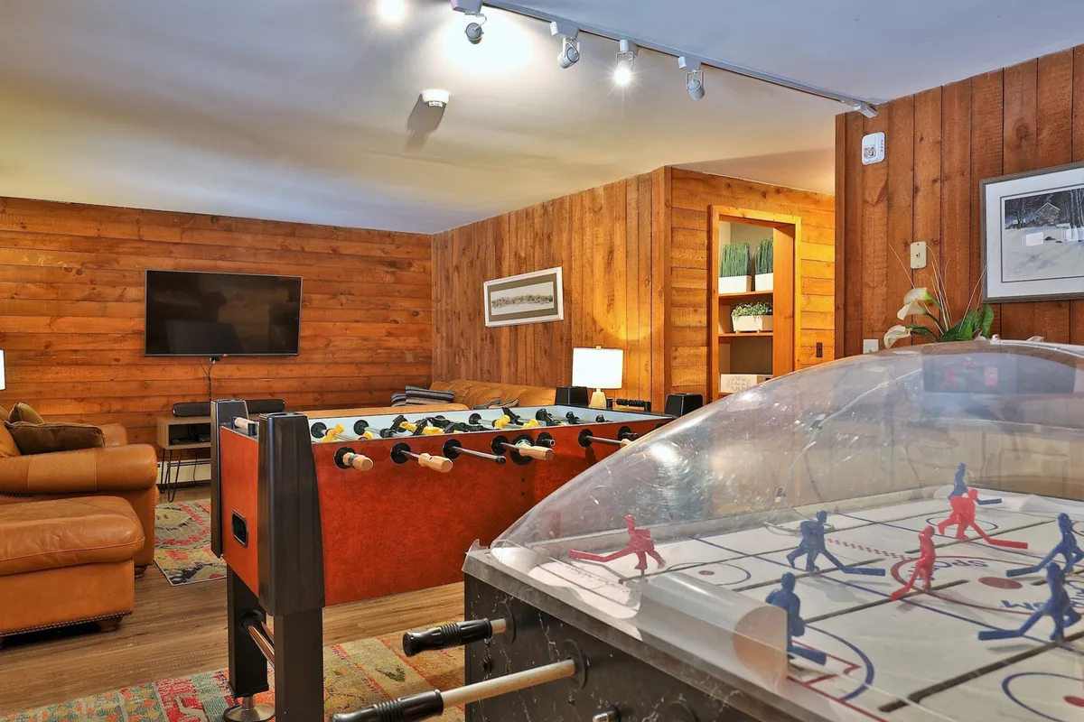 A vibrant game room featuring a foosball table, air hockey table, comfortable seating, and wooden wall paneling. The mood is fun and casual—perfect for group entertainment after skiing.