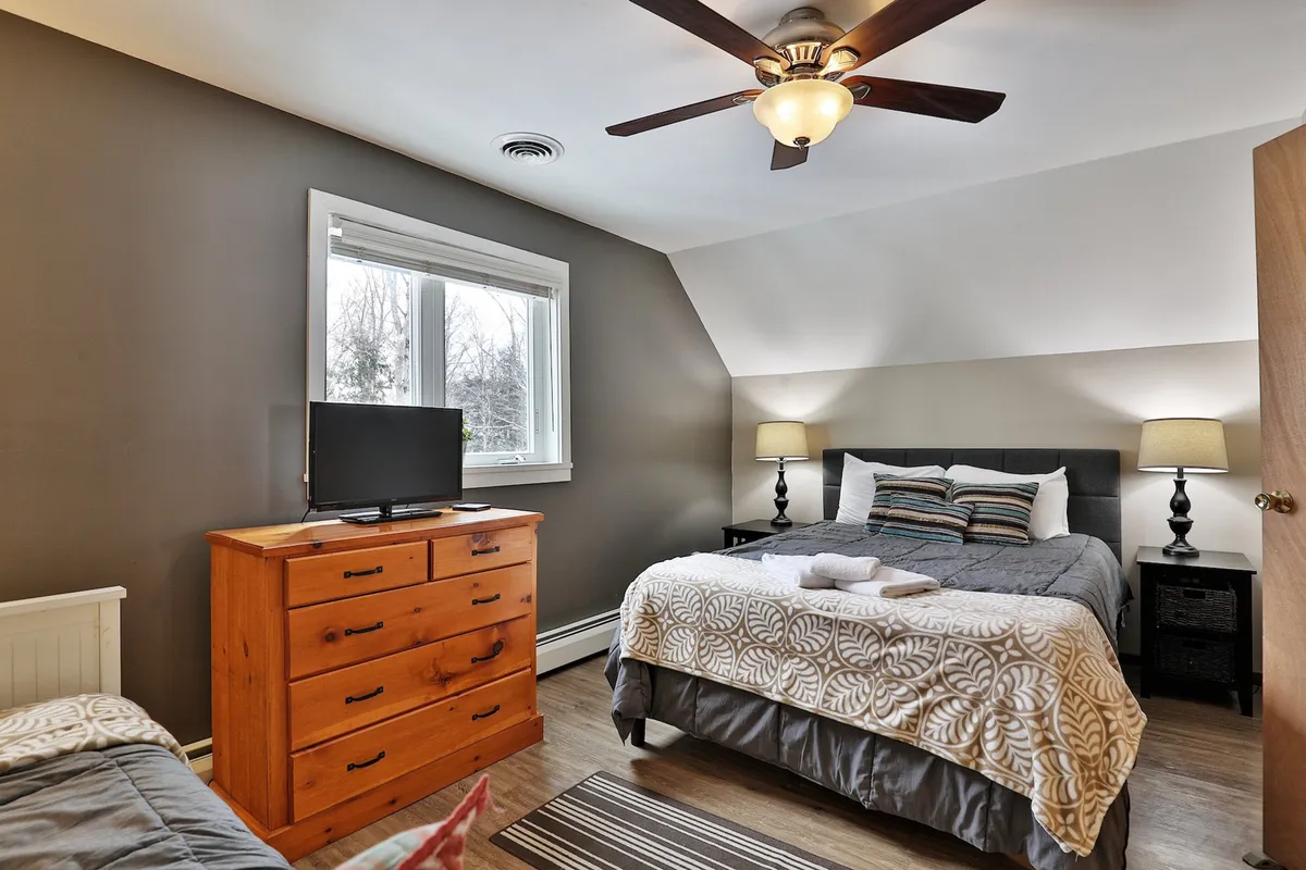 A sunlit bedroom with a large bed featuring a decorative throw, a dresser with a TV on top, matching bedside lamps, and a big window that fills the room with natural light. The overall mood is restful and inviting.