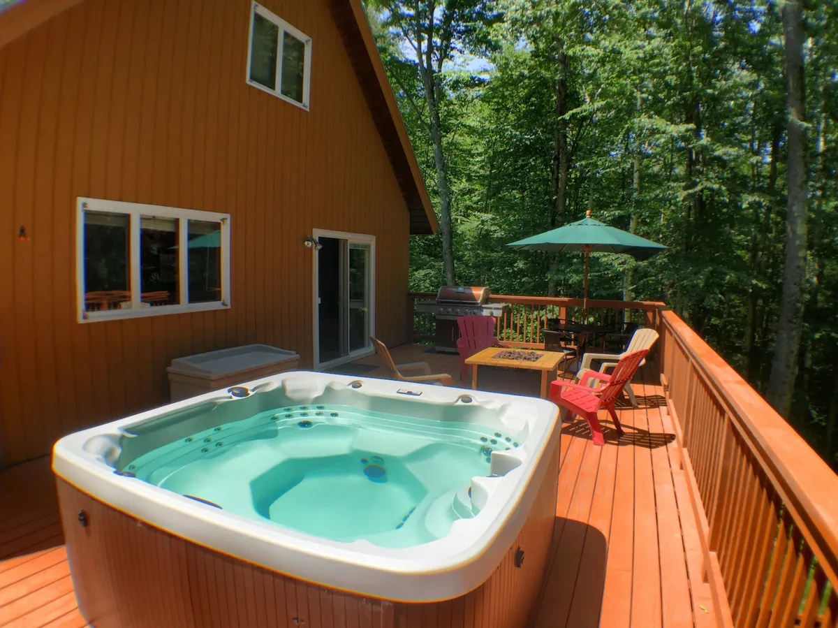 An inviting exterior deck features a bubbling hot tub, ample seating, a dining table, and a grill. Surrounded by trees, the space is perfect for evening relaxation or group gatherings under the stars.