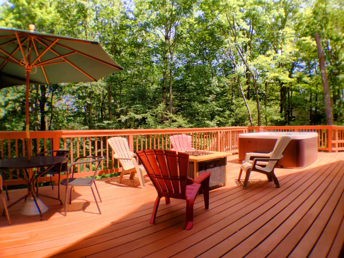 The image displays adirondack chairs, an outdoor dining table, a hot tub, and lush greenery surrounding the deck. The setting is designed for relaxation, outdoor meals, and enjoying nature—activities that align with summer recreation and family bonding.