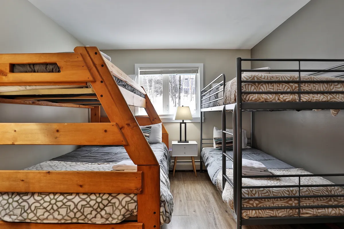 A cozy bunk room featuring sturdy wooden bunk beds illuminated by a soft lamp, with a window letting in natural light. The room has an inviting, playful ambiance, ideal for kids or groups seeking a fun, communal sleeping space.
