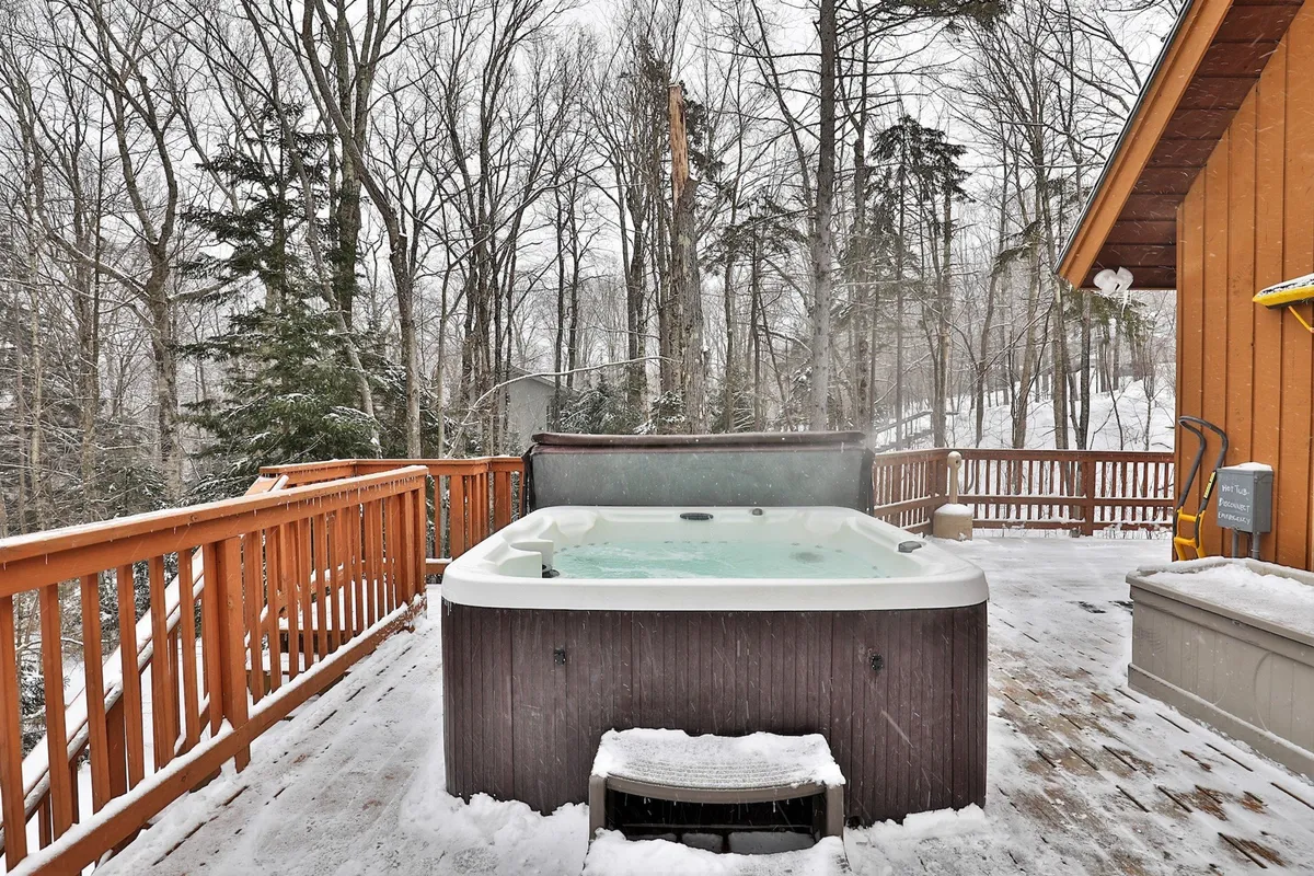 A steaming hot tub is set on a deck surrounded by trees and a snowy landscape. The setting is tranquil and cozy, offering a perfect spot for relaxation after a day of skiing or snowboarding.