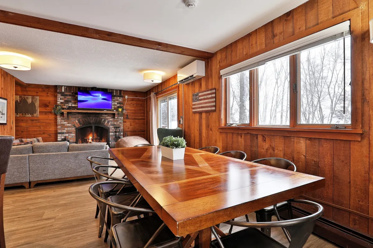 An inviting open-plan dining and living area featuring a dining table, cozy sofa, fireplace, television, and expansive windows letting in natural light. The space seamlessly supports a range of group activities from meals to relaxing by the fire.