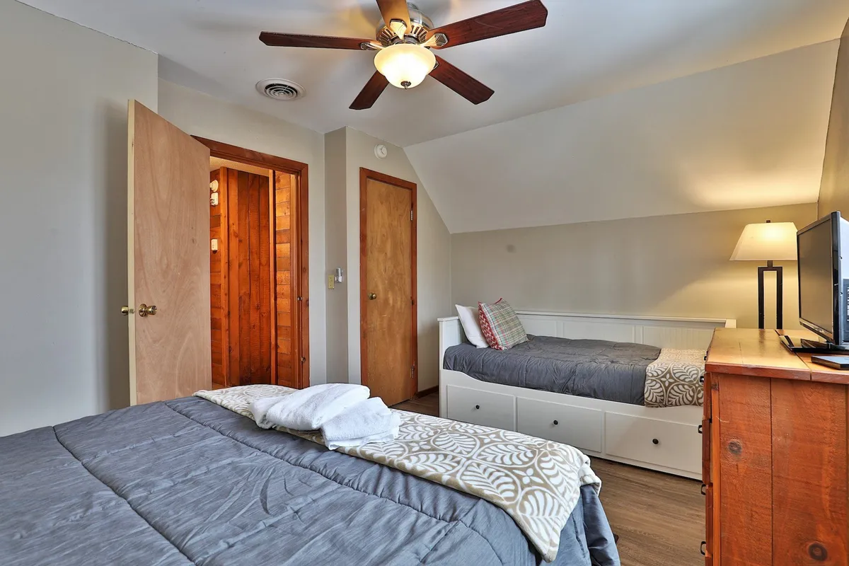 A spacious bedroom with a queen bed dressed in gray bedding and a single bed adorned with decorative pillows. A wooden dresser, bedside lamp, and wood-paneled doors give the space a warm, comfortable vibe—showcasing flexible sleeping arrangements for guests.