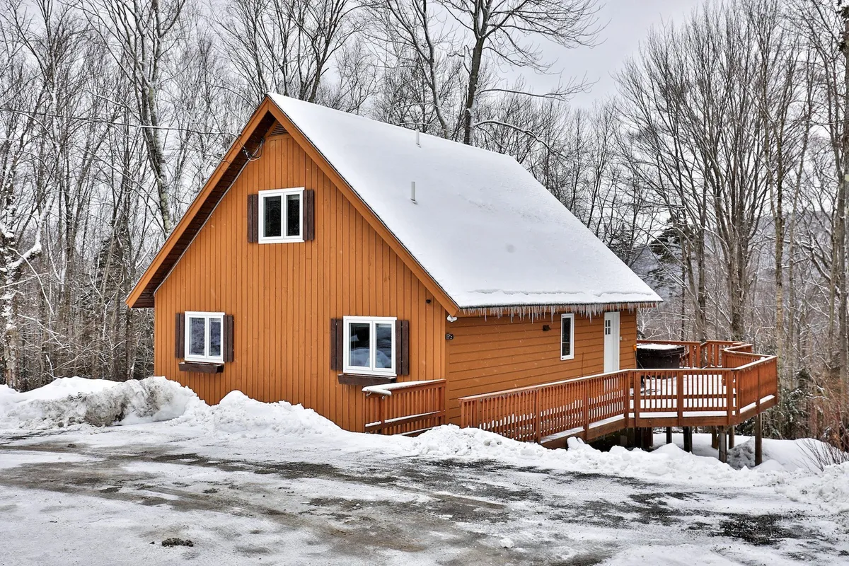 This exterior image features a charming wooden A-frame structure with a snow-covered roof, a spacious deck area, and surrounding trees. The scene evokes the quintessential Vermont winter getaway, with the home nestled in a peaceful, snowy landscape.