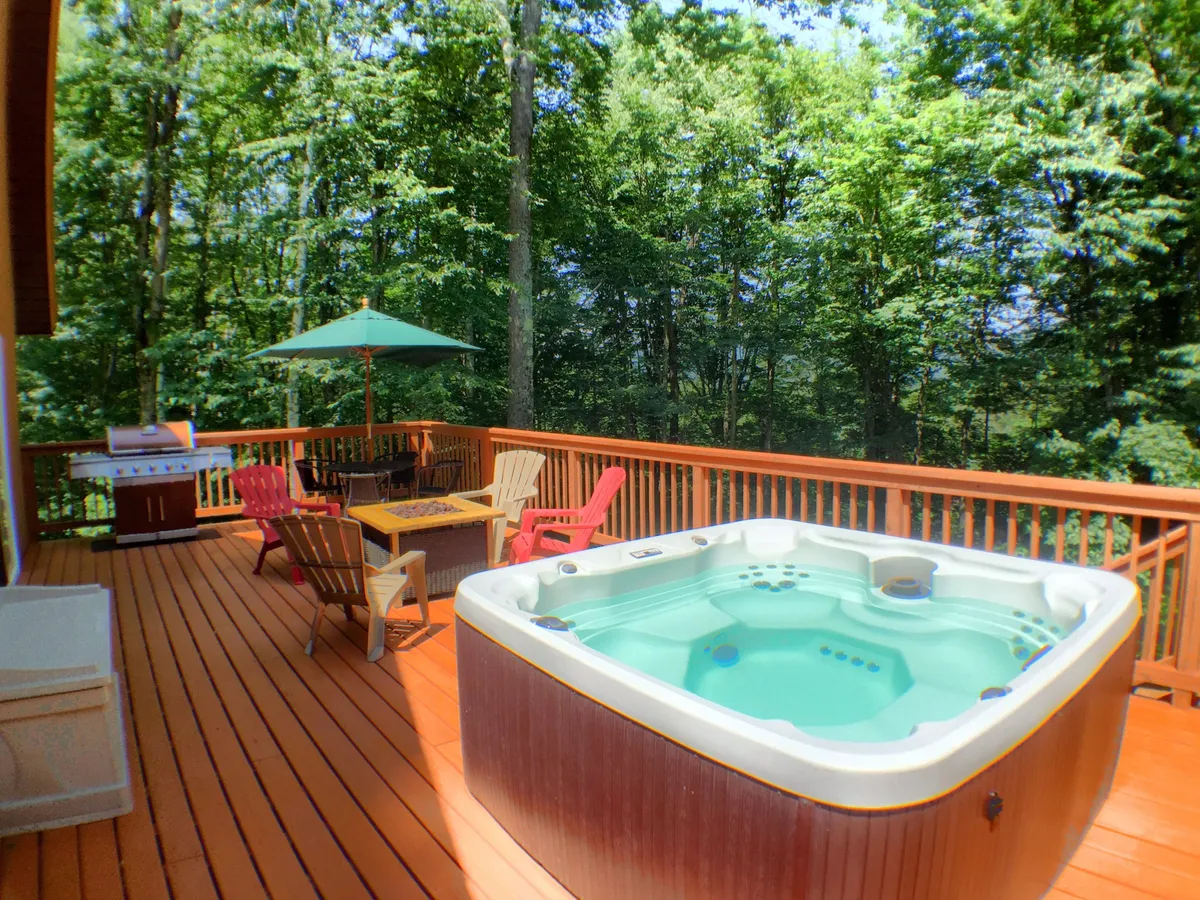The image showcases the inviting exterior of a spacious wooden deck set against a scenic Vermont backdrop. Prominently featured are a bubbling hot tub, ample outdoor seating with an umbrella, a grill, and space for social gatherings—all surrounded by nature, perfect for après-ski relaxation or stargazing evenings.
